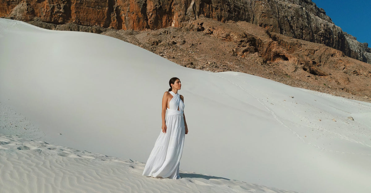 Woman wearing a flowing white Bohème by Ena dress on white sand dunes with rocky cliffs in the background