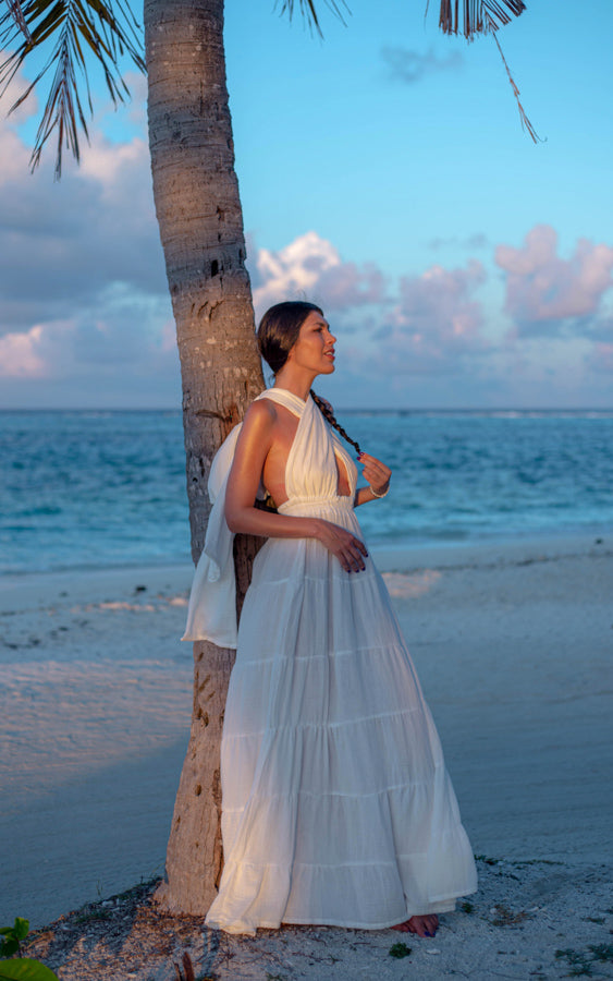 Woman wearing the white Esmeralda Versatile Maxi Dress beside a palm tree by the sea