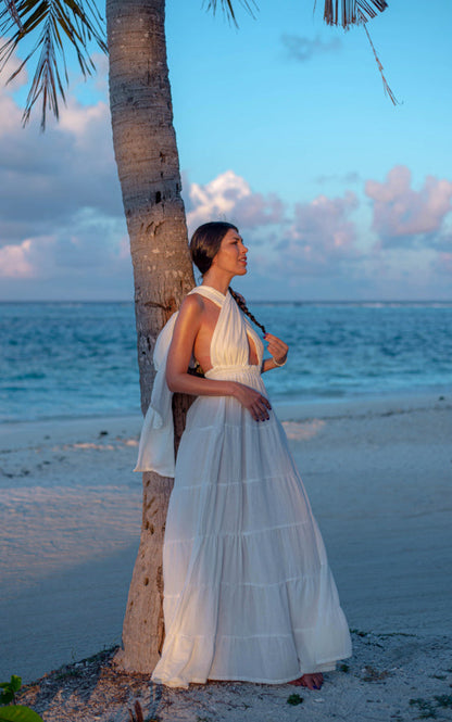 Woman wearing the white Esmeralda Versatile Maxi Dress beside a palm tree by the sea